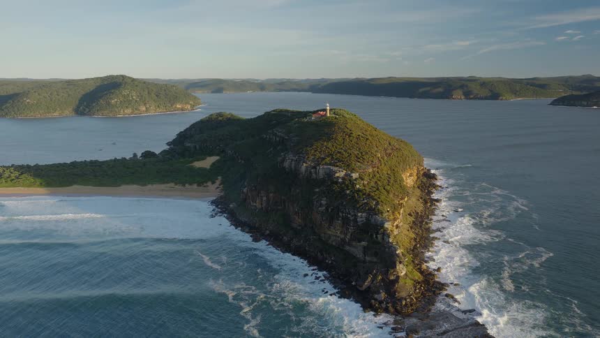 An aerial view of waves crashing against a large rugged rock formation topped with a lighthouse situated in a scenic bay with lush green hills in the background