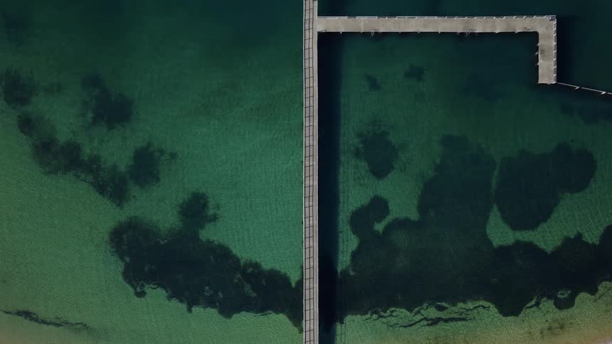 An aerial view shows a long pier stretching out into a turquoisegreen sea the water near the shore is lighter and sandy while deeper patches of dark green suggest underwater reefs or plant life