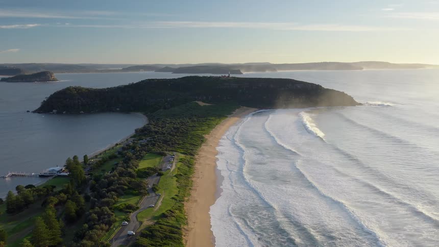 An aerial view of a coastline shows a thin tree covered strip of land jutting out into a bay with a sandy beach and waves to its right and a calm body of water with a white pier to its left