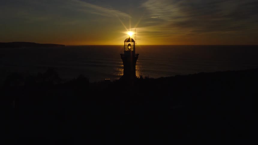 Lighthouse overlooking the water under a dark sky as the sun on the horizon reflects on the ripples and waves