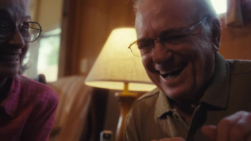 Elderly man making gestures with his hands and laughing together with an elderly woman indoors during day time