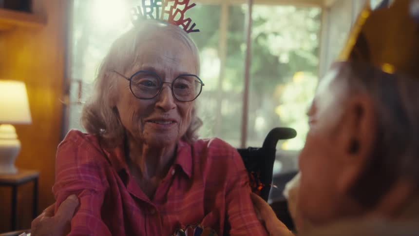 An elderly woman is sitting in a wheelchair wearing a happy birthday headband and glasses she is smiling and looking at another person offscreen as if she is being celebrated