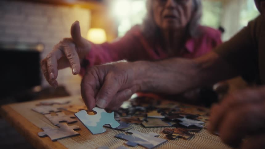 Close up of an elderly man and woman arranging a jigsaw puzzle on a table in a living room during daytime