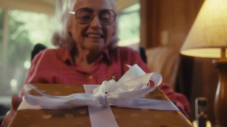 Elderly woman smiling and opening a birthday gift while elderly male hands gesticulate next to her indoors during day time