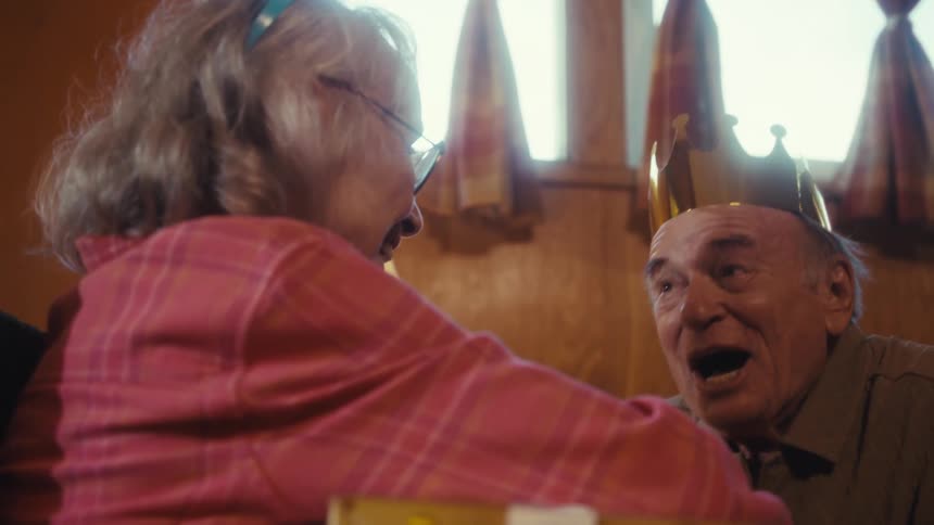 Elderly man and woman wear party hats while exchanging a gift at home during the daytime