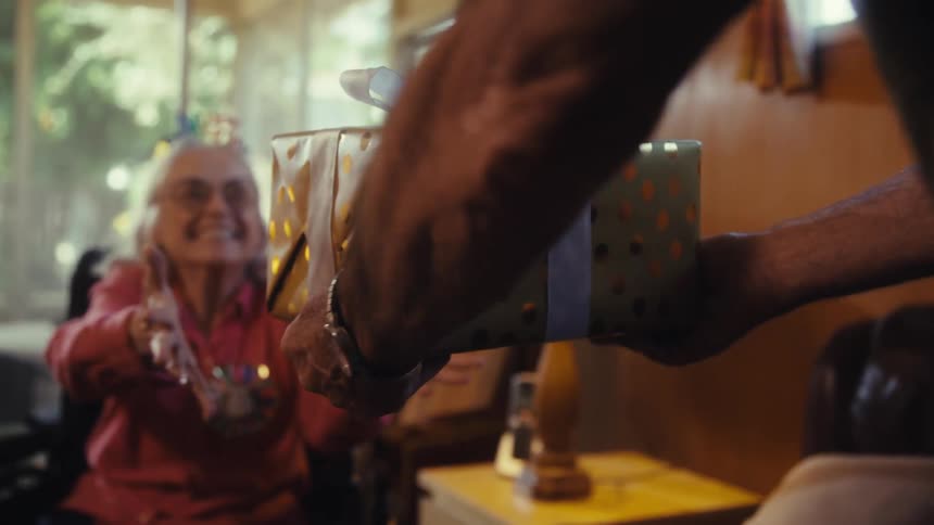 A delighted grandma reaches for a wrapped present laughing as sunlight filters through the nearby window