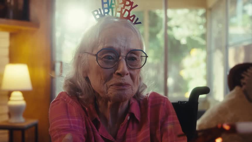 A senior woman in a wheelchair wearing a happy birthday headband and my big day badge is laughing happily while talking to a man