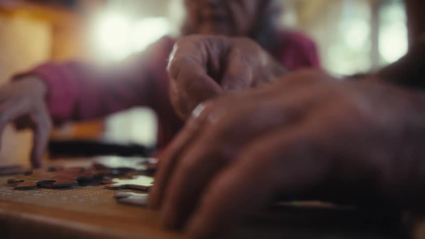 The hands of two people are seen in a closeup shot as they collaboratively work on a wooden tabletop puzzle