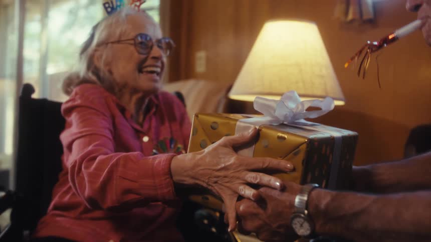 A man presents a birthday present to an elderly woman sitting in a living room on her birthday