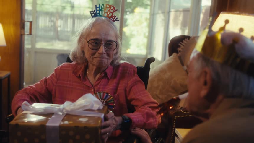 Elderly man wearing a party hat hands a present to an elderly woman wearing a birthday hat and they hug indoors during day time