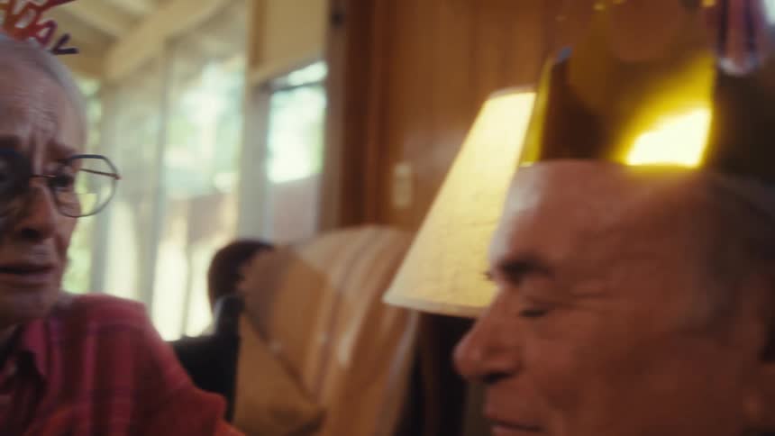 Elderly woman with glasses and birthday tiara smiles excitedly as she talks to an elderly man