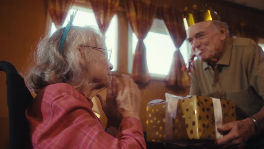 An elderly woman in a plaid shirt and a happy birthday headband initially engrossed in her phone is surprised by her husband who approaches with a wrapped gift and they share a loving embrace