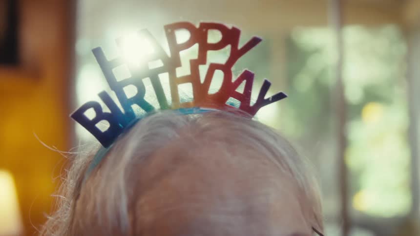 Elderly woman with a birthday tiara sitting talking to another person in the room