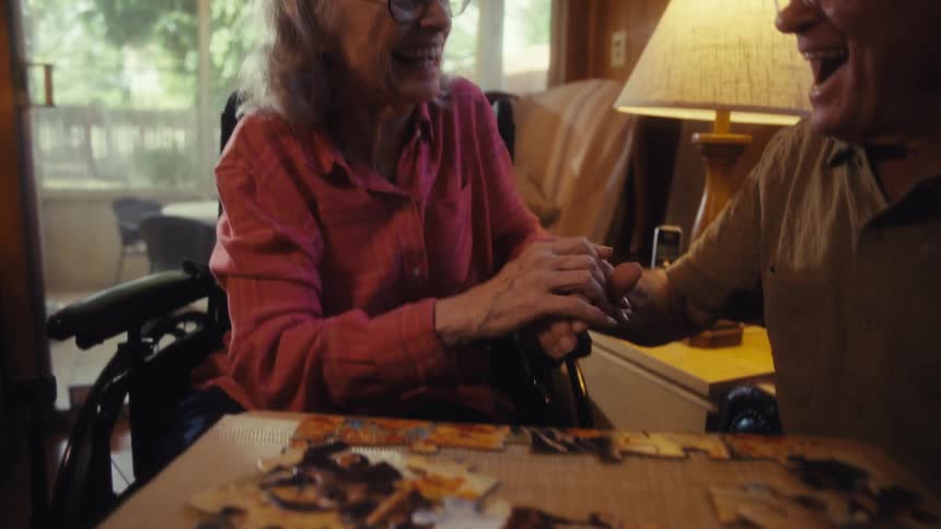 Elderly man and woman sitting indoors playing with a jigsaw people while laughing and celebrating by holding hands during day time