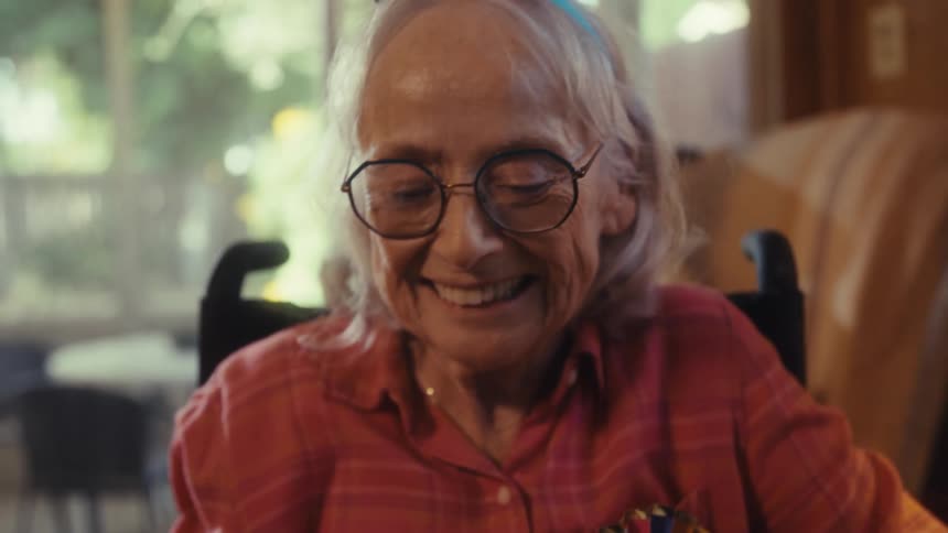 Elderly woman wearing a happy birthday headband covers her mouth emotionally