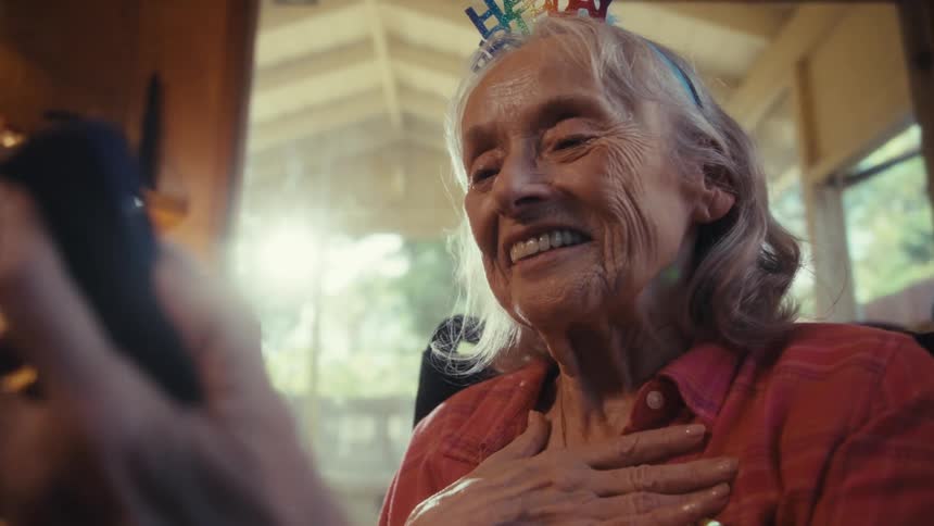 Happy grandmother videochats on her birthday hand to chest in warm window light