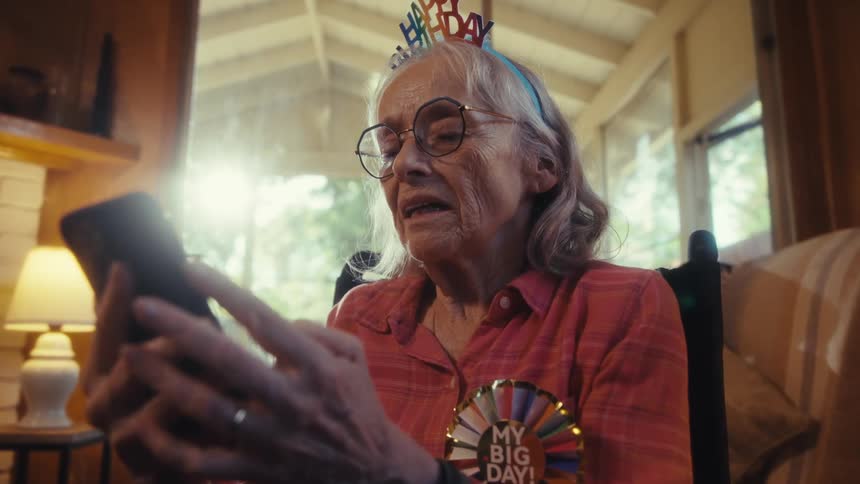 An elderly woman sitting in the living room holding and scrolling on a mobile phone while smiling