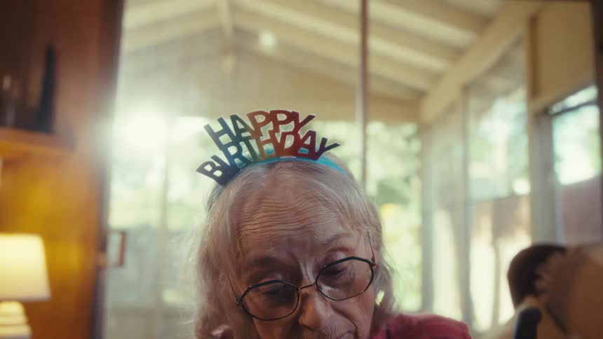 An elderly couple celebrating a birthday with the woman wearing a happy birthday hat and glasses holding a present while the man stands by her side