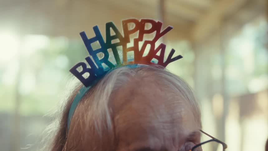 An elderly couple in the living room the woman sitting in a wheelchair wearing a birthday hat and holding a cake talking to the man sitting on a couch both filled with excitement
