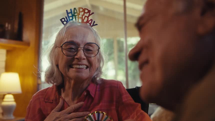 Elderly woman smiling with a birthday headband while receiving affection on her face