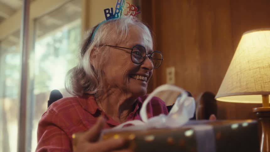 An elderly woman in a wheelchair joyfully celebrates her birthday reacting with surprise happily receiving a gift and sharing a warm hug with a loved one