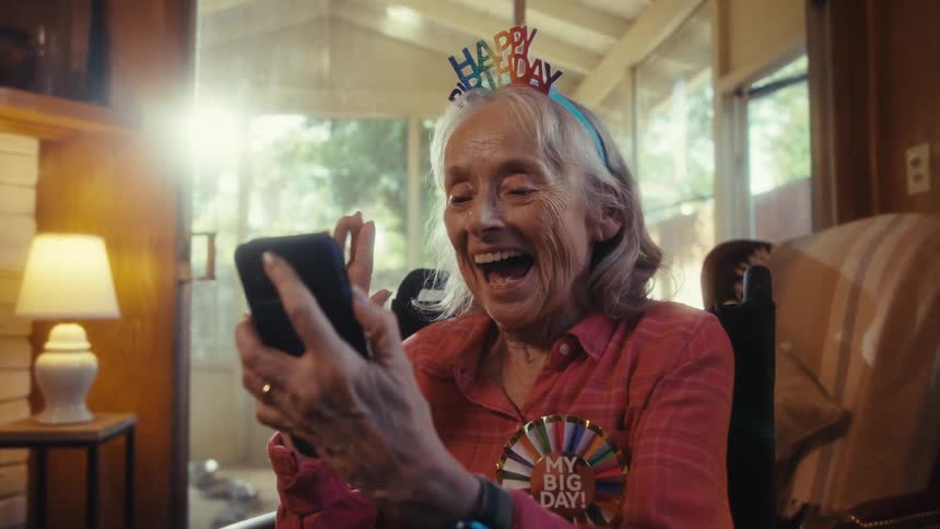 Elderly woman wearing a party hat sitting down on a wheeling chair using her smart phone while waving and smiling at it indoors during day time