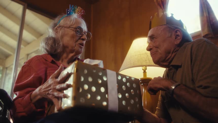 The video shows an older woman wearing a happy birthday headband holding a large wrapped gift she is giving the present to an older man who is also wearing a birthday crown the two are sitting indoors with a lamp and curtains visible in the backgr