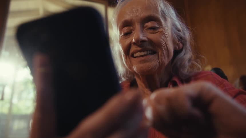 An elderly woman smiles at a smartphone indoors