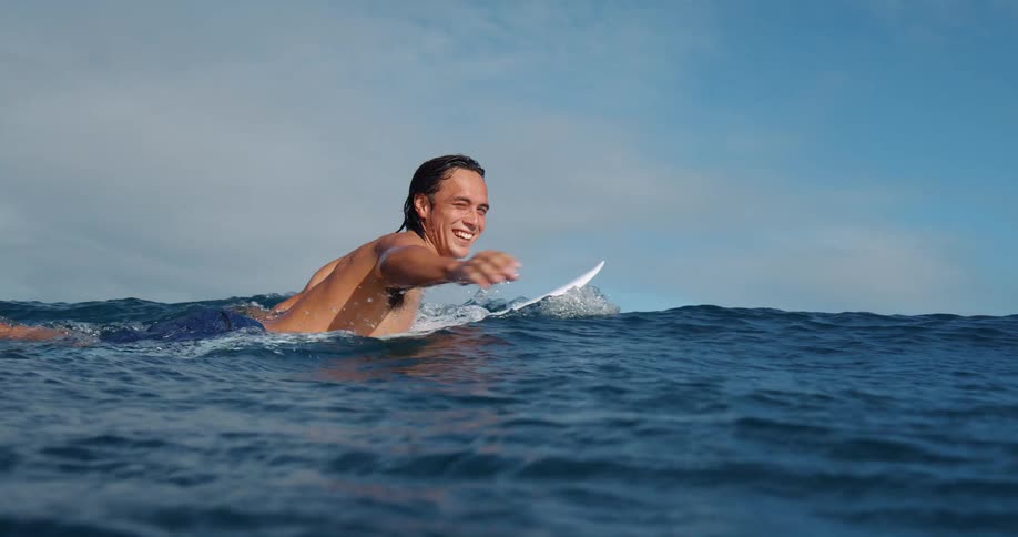 A smiling man paddles a surfboard in the ocean