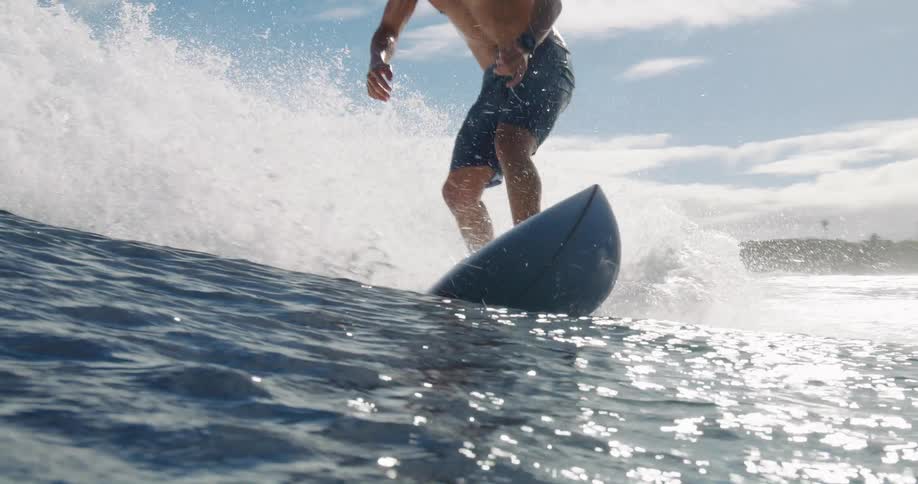 A person is seen from a low angle successfully riding and balancing on a surfboard as it moves through a large powerful ocean wave on a sunny day