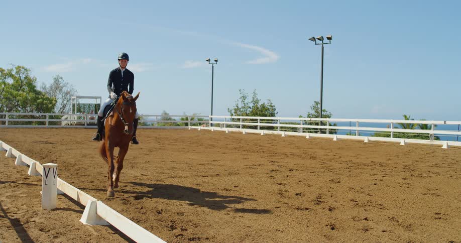 A woman rides a brown horse around an enclosure surrounded by white fences