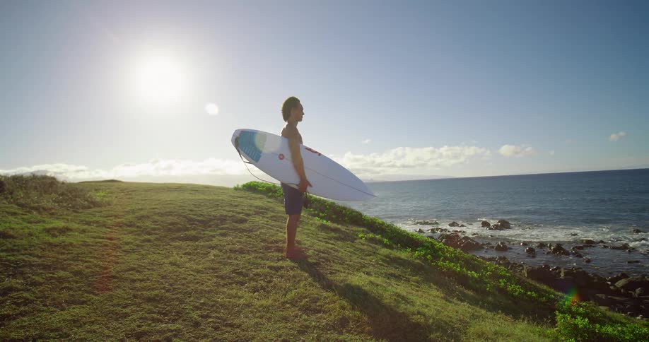 A surfer holding his board stands on a grassy hill overlooking the ocean on a bright sunny day as the camera slowly pushes in on him
