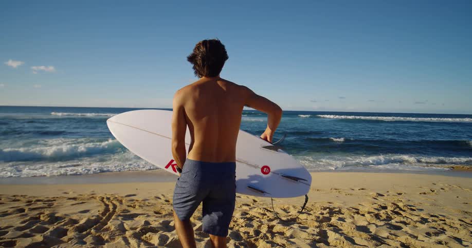 A man holding a surfboard is walking toward the ocean on a sunny beach ready to surf