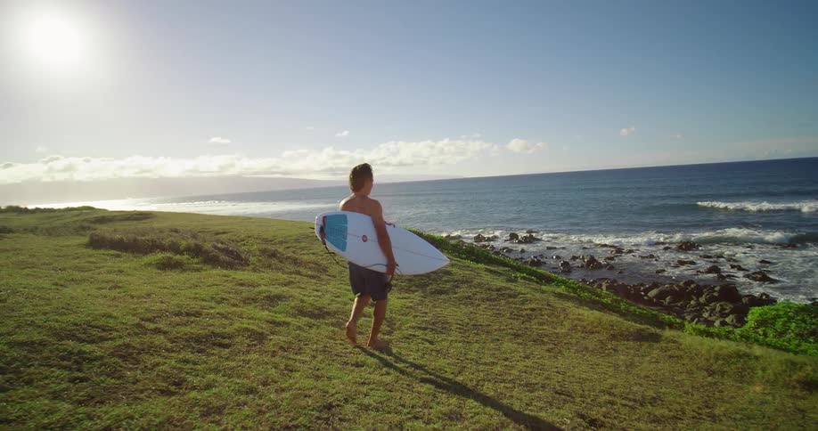 Man carries his surfboard down a grass covered hill as waves roll into shore and splash against rocks along the coast