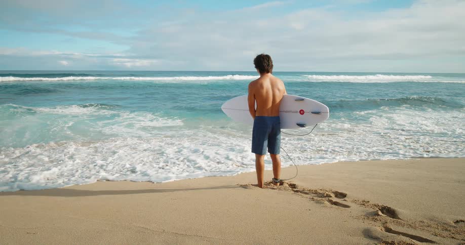 White man holding a surf board and standing on a beach looking at the sea and the horizon outdoors during day time