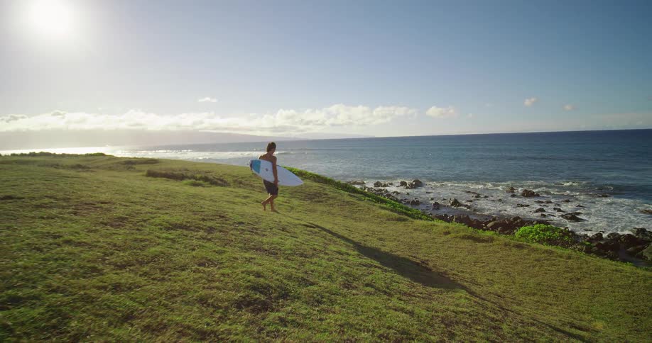 A person runs with a surfboard under their arm on a green hill next to a rocky ocean coast