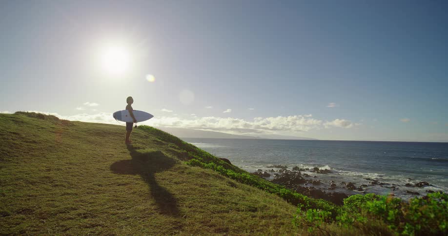 A man stands on a hill overlooking the ocean while holding a surfboard
