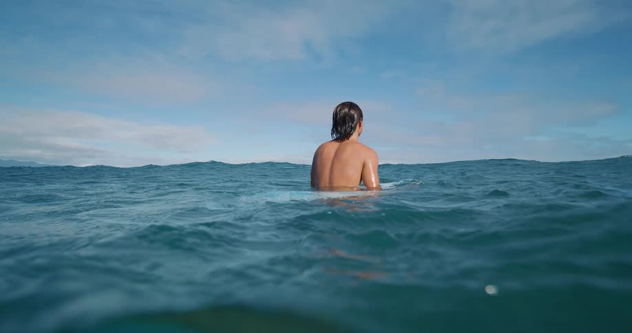 Young male surfer sitting down floating on a surfboard in the middle of the sea outdoors during day time