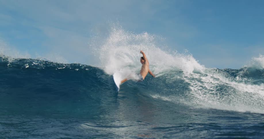 A shirtless surfer in blue shorts rides a large blue ocean wave on a white surfboard spraying water as the wave breaks