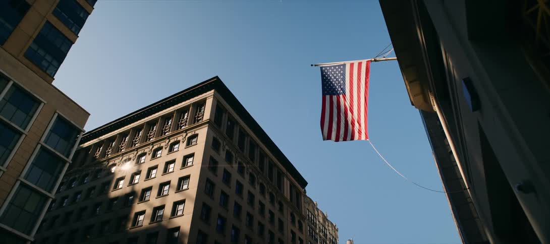 An american flag is hanging vertically from a building on a city street with the sun shining and reflecting off the windows of a large classic building in the background
