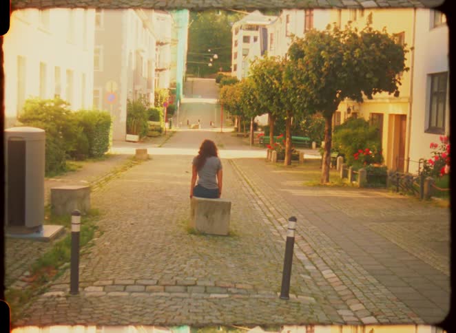 Woman sitting on a stone block looks at a cobblestone street lined with trees and houses
