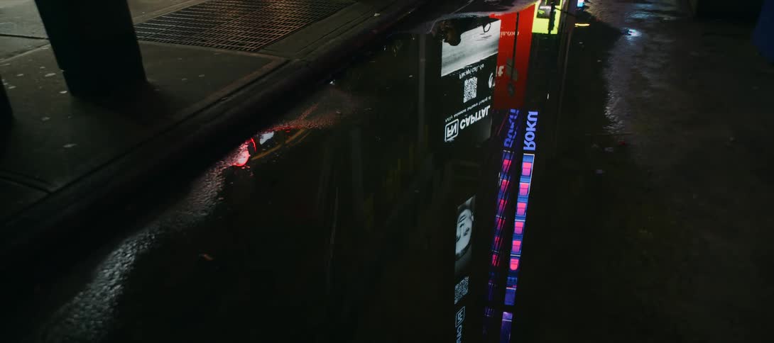 The colorful lights of city advertisements reflect in a dark puddle on a wet street at night as a persons foot walks by on the curb