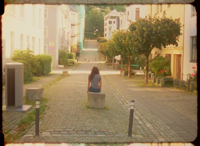 Person sitting alone on a stone bench in a quiet street with trees and houses