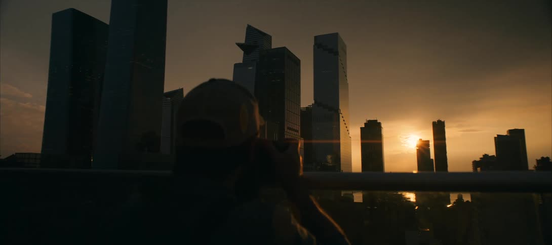 A silhouetted person looks at a cityscape at sunset with tall dark skyscrapers visible as the sun hangs low behind them casting an orange glow across the sky