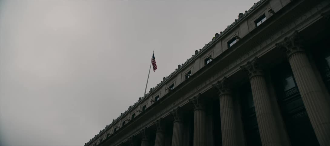 A lowangle shot shows an american flag flying above an imposing government building with large columns under a grey overcast sky