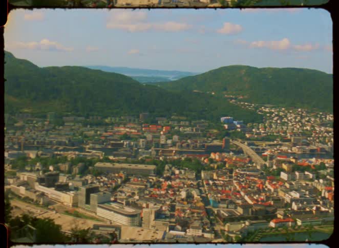 The video shows a beautiful wideangle shot of a city nestled between mountains and a bay with buildings scattered up the hillsides the camera pans slowly down to reveal people enjoying the scenic view from an observation deck