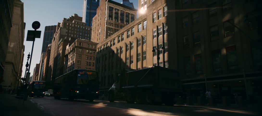 Afternoon light cuts through tall buildings as vehicles move along a busy street