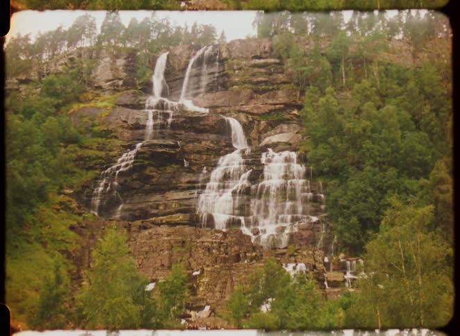 An old film camera shows a large tiered waterfall flowing down a rocky tree covered cliff with several people at the bottom