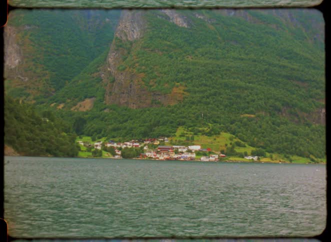 Water flows past a coastal town surrounded by forested mountains during the daytime