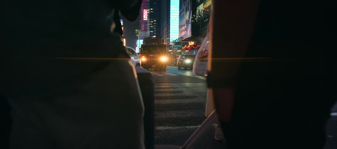 Headlights roll toward a busy crosswalk framed by nighttime city glow